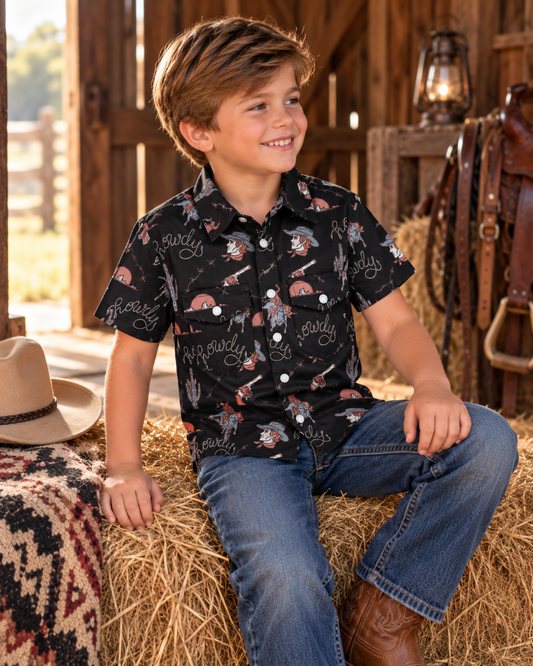 A young boy wearing a black short-sleeve button-up shirt with a cowboy and cattle print, blue jeans, and brown boots, sitting on a hay bale in a rustic barn setting.