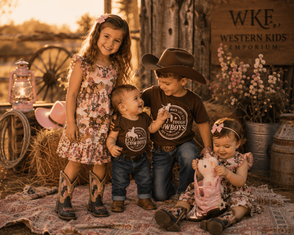 Four children posing together in a rustic Western-themed studio setting. Two children wear matching brown 'Western Kids Cowboys' t-shirts, while the other two wear floral dresses.