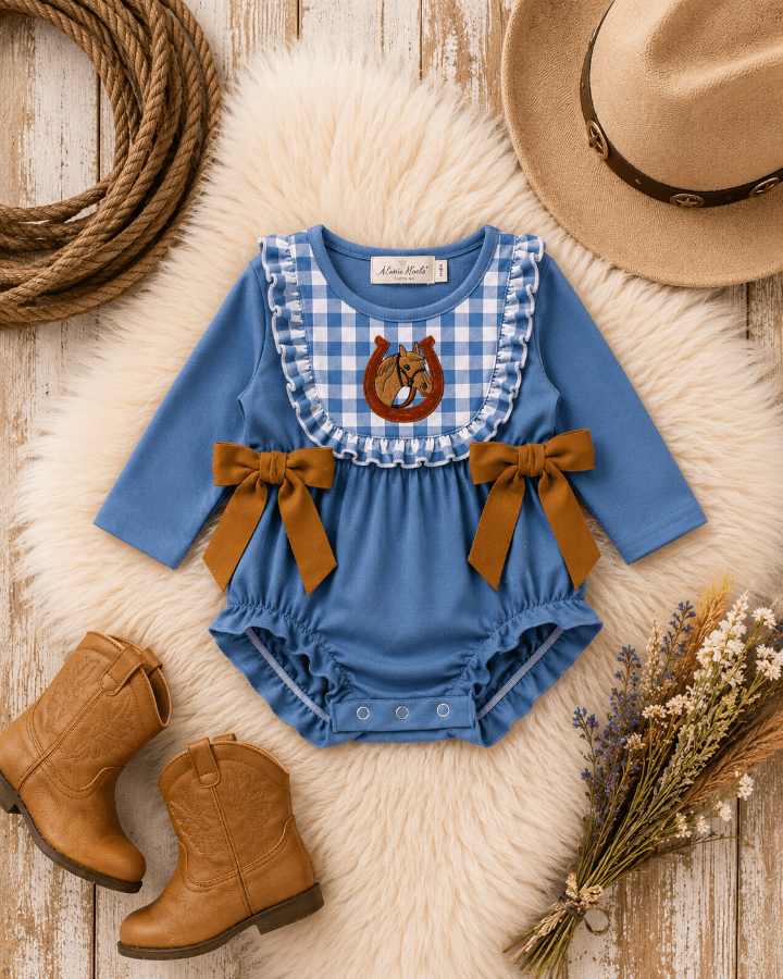 A baby girl's long-sleeve romper in dusty blue, styled in a rustic flat-lay with a faux fur rug, cowboy boots, a rope, a straw hat, and dried flowers. The romper features a blue and white gingham bib panel with a brown horseshoe graphic, and two brown fabric bow accents on the sides.