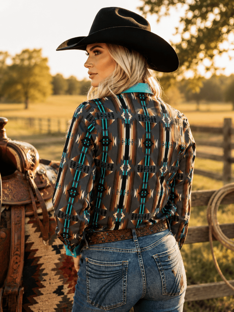 A woman wearing a black cowboy hat, a long-sleeve button-up shirt with a brown, black, and turquoise geometric pattern, and blue jeans, standing in a sunny outdoor ranch setting next to a leather saddle.