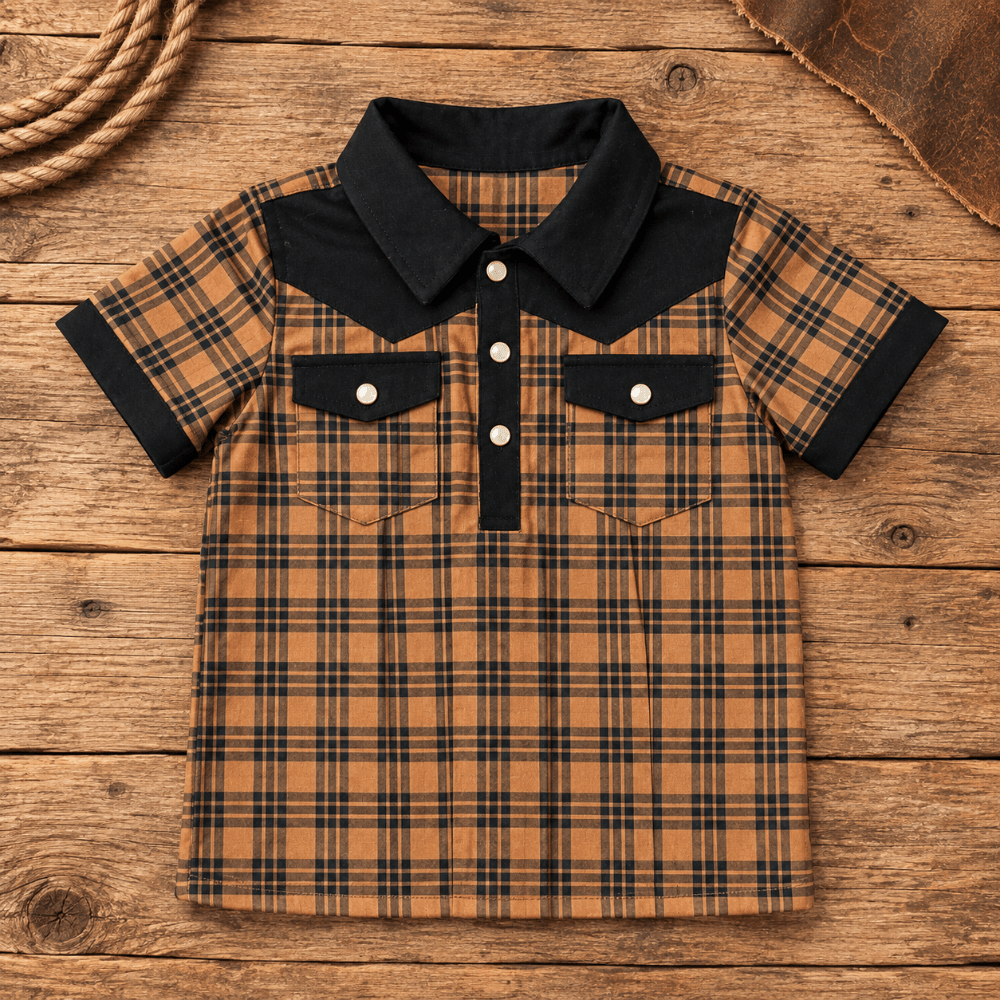 A short-sleeve plaid shirt for kids, featuring a black collar, cuffs, and button placket, with a brown and black plaid body. The shirt is laid flat on a rustic wooden background with decorative rope and leather in the corners.