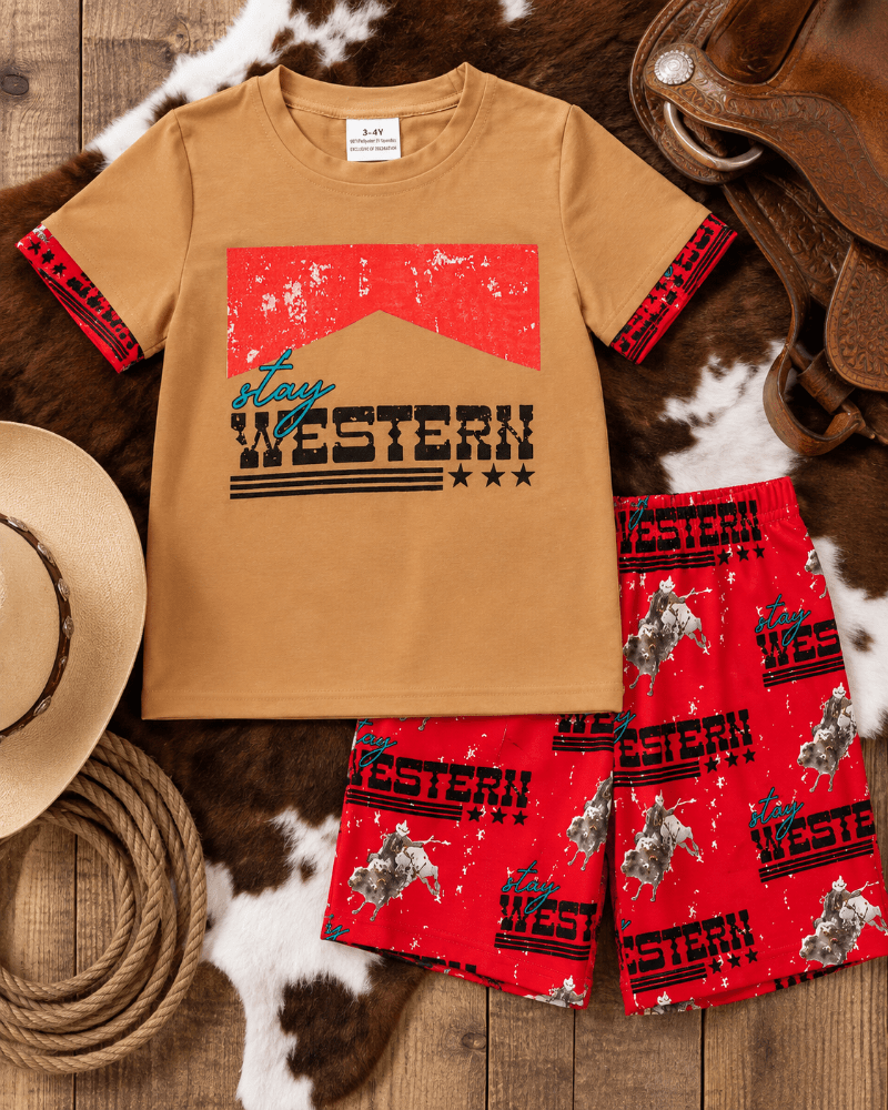 A children's two-piece western-themed outfit laid flat on a cowhide rug with rustic wood and western props. The set includes a tan short-sleeve T-shirt with red and black sleeve trim and a large distressed 'Stay Western' graphic on the front, plus matching red shorts covered in a western rider print and 'WESTERN' text.