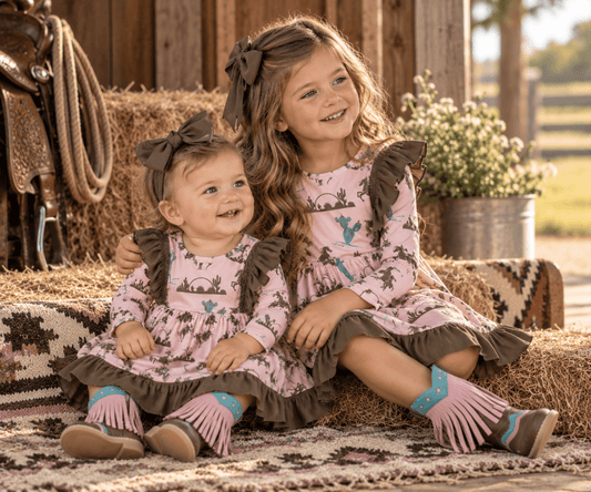 Two young girls sitting together on a hay bale in a rustic barn setting, wearing matching pink long-sleeve dresses with a brown cactus print, brown ruffled shoulder details, and brown bows in their hair.