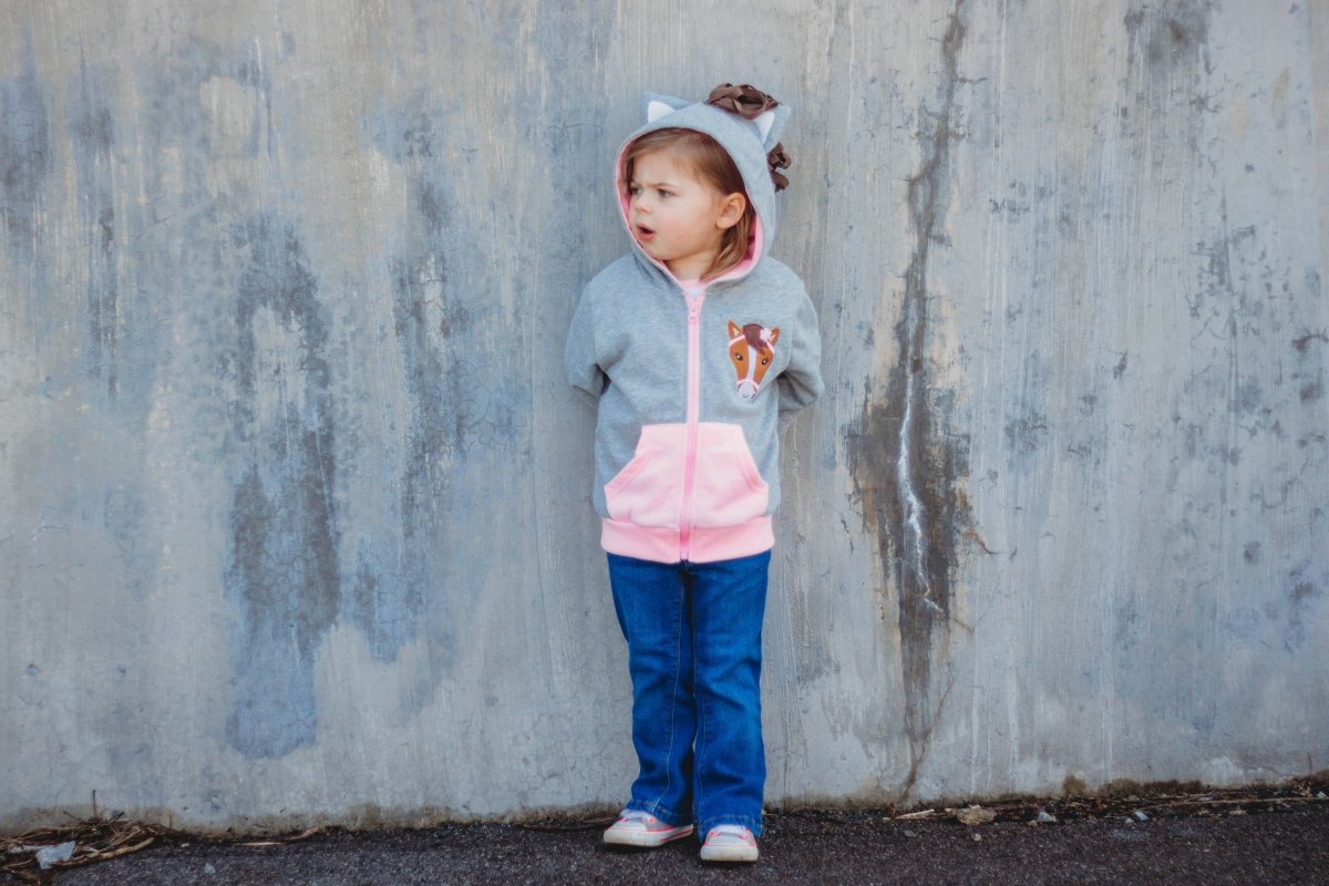 Child wearing a gray and pink hoodie with a horse design against a textured wall.
