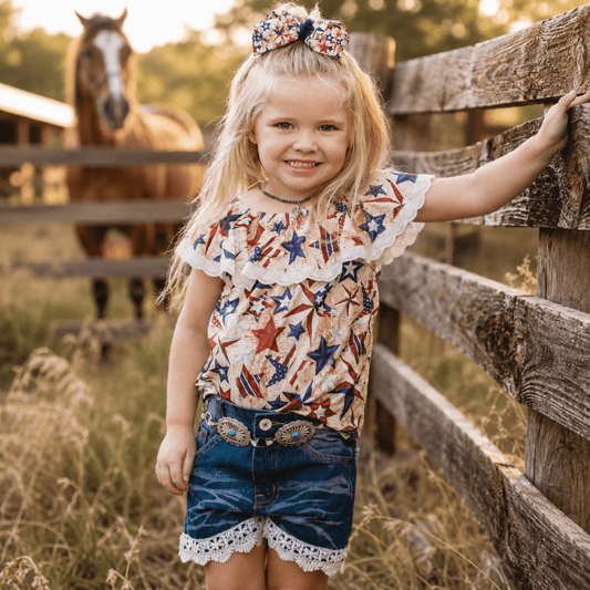 A young girl stands outdoors by a rustic wooden fence in a field, wearing a patriotic red, white, and blue star-patterned top with ruffled sleeves and denim shorts. A horse is visible in the background behind the fence.