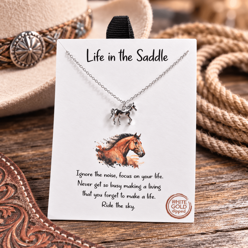 A silver horse pendant necklace on a black display card with the words 'Life in the Saddle' and a western-themed background with a hat, rope, and wooden surface.