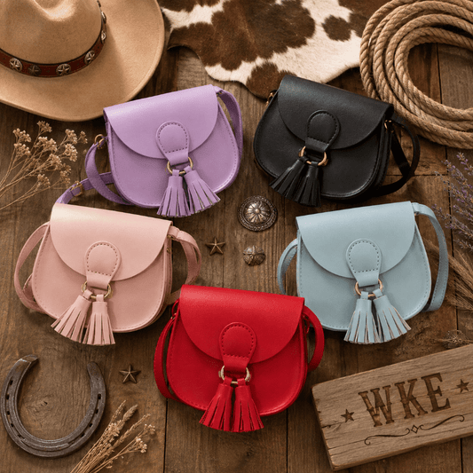 Five faux leather saddle crossbody bags in pastel purple, pink, light blue, and red, arranged on a rustic wooden surface with western-themed props including a hat, rope, horseshoe, and wooden sign.