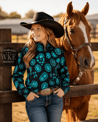 A woman in a black cowboy hat and a black long-sleeve quarter-zip shirt with a bright turquoise floral pattern stands next to a brown horse in front of a wooden fence. The sign on the fence reads 'WESTERN KIDS WKE EMPORIUM'.
