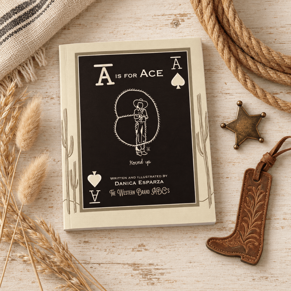 A Western-themed notebook or book cover styled in a flat lay on a light wood surface, surrounded by a lasso, burlap, wheat stalks, a brass star, and a leather cowboy boot keychain. The cover is black with cream lettering and illustrations, including the text 'A IS FOR ACE' and 'The Western Band P.
