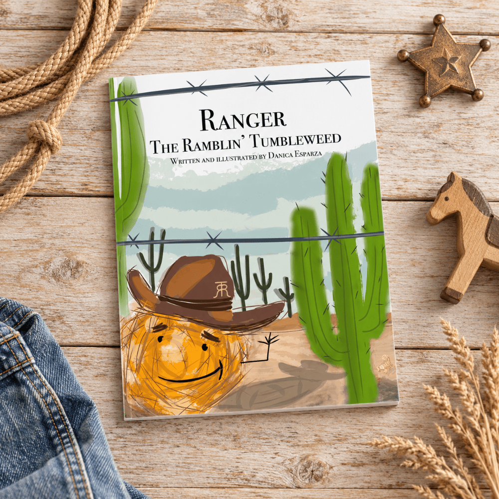 A children's book titled 'Ranger The Ramlin' Tumbleweed' lies on a rustic wooden surface, surrounded by western-themed props including a rope lasso, denim fabric, a wooden toy horse, a star badge, and dried wheat. The cover shows a desert scene with cacti, barbed wire, and a smiling tumbleweed wearing a cowboy hat.