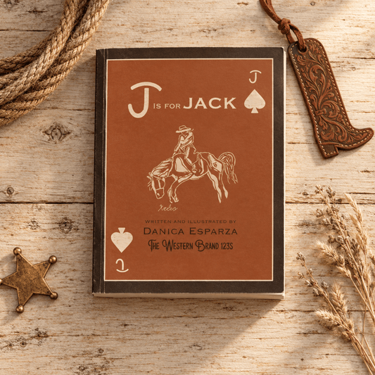A rectangular book or notebook titled "J is for Jack" lies on a rustic wooden surface, surrounded by western-themed props including a rope, a leather boot-shaped tag, a brass star, and dried grass. The cover is brown with cream lettering and illustration of a rider on a horse.