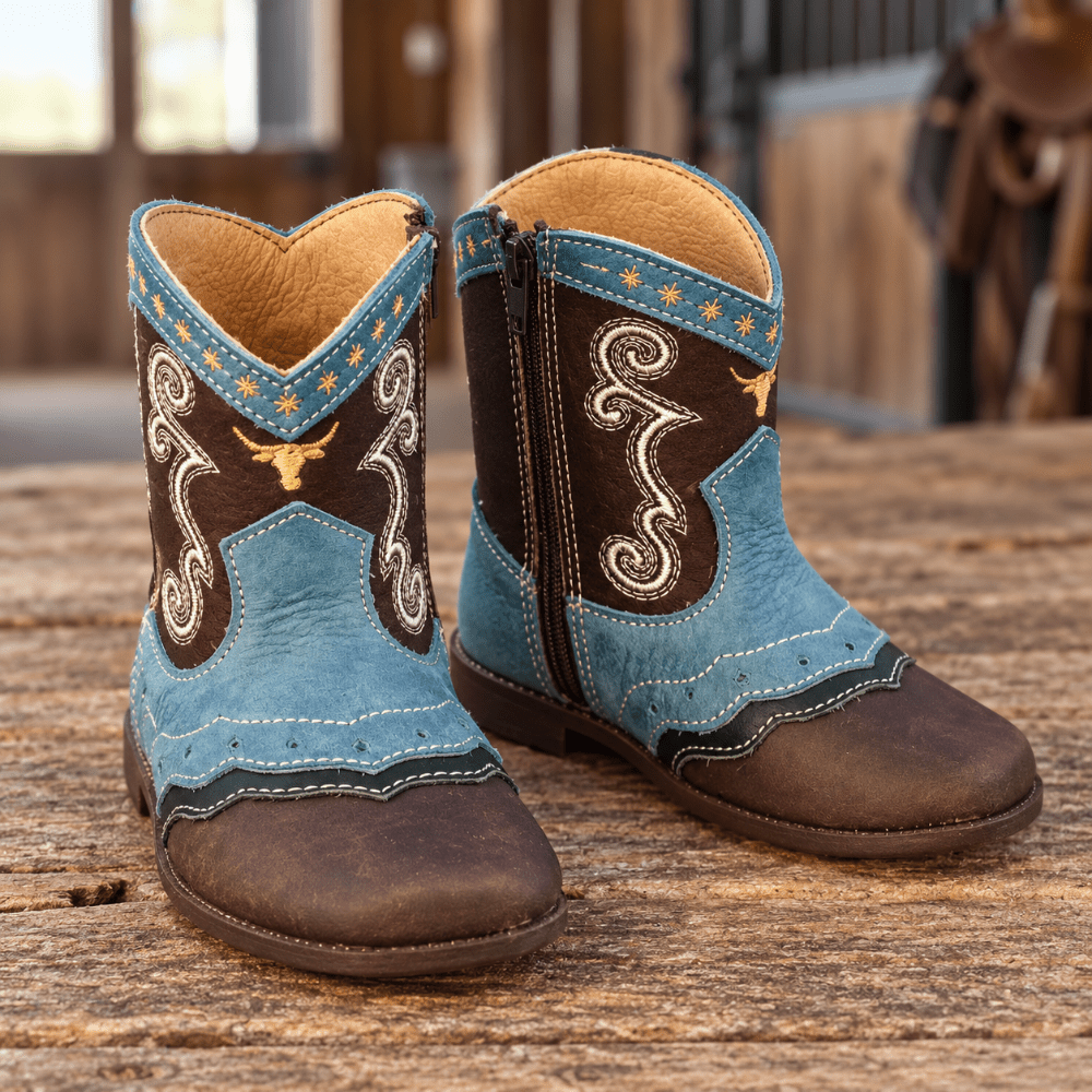 A pair of western-style cowboy boots with light blue and dark brown panels, white embroidery, and gold star accents, displayed on a rustic wooden surface in front of a blurred barn interior.