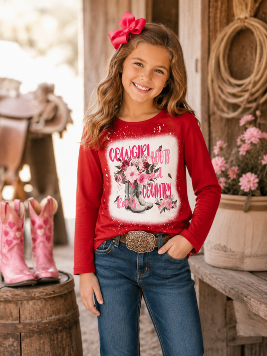 Young girl in a red shirt with a cowboy graphic, standing in a rustic setting with pink boots and flowers.