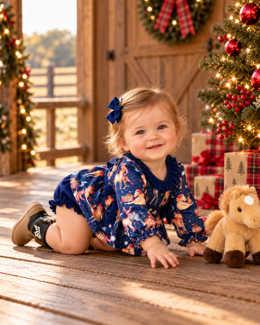A toddler girl wearing a navy blue Christmas-themed long-sleeve romper with a deer print and a matching hair bow, crawling on a wooden porch next to a plush toy horse with a Christmas tree in the background.