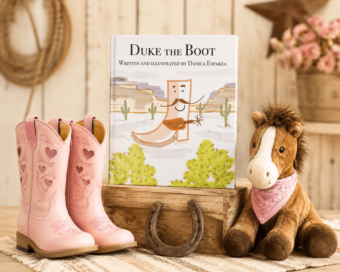 A children's book titled 'Duke the Boot' stands on a wooden crate between a pair of pink western boots with heart details and a plush toy horse wearing a pink bandana. The scene is styled with rustic decor, including a horseshoe and rope, against a light wood-paneled background.