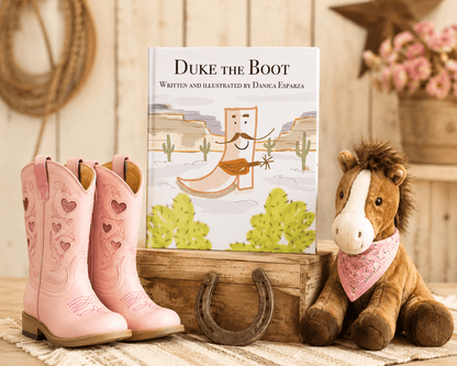 A children's book titled 'Duke the Boot' stands on a wooden crate between a pair of pink western boots with heart details and a plush toy horse wearing a pink bandana. The scene is styled with rustic decor, including a horseshoe and rope, against a light wood-paneled background.