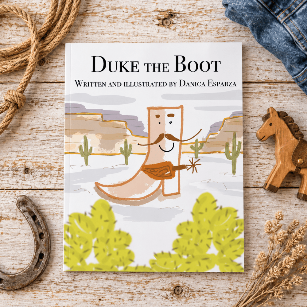 A children's book titled "Duke the Boot" by Danica Esparza lies on a rustic whitewashed wooden surface, surrounded by western-themed props including a rope, a wooden toy horse, a horseshoe, and dried grass. The cover shows a cowboy boot character playing a guitar in a desert landscape.