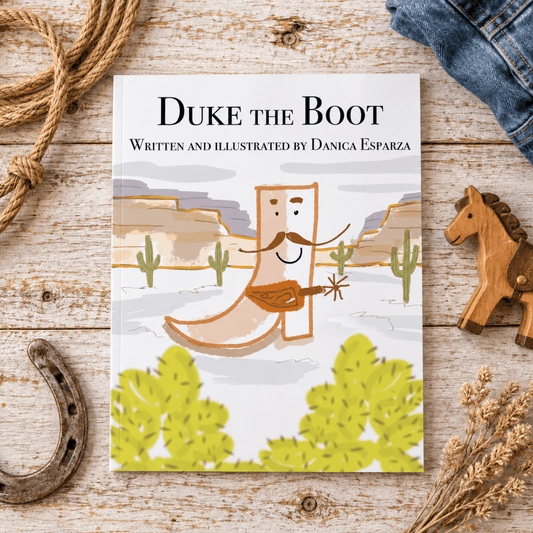 A children's book titled "Duke the Boot" by Danica Esparza lies on a rustic whitewashed wooden surface, surrounded by western-themed props including a rope, a wooden toy horse, a horseshoe, and dried grass. The cover shows a cowboy boot character playing a guitar in a desert landscape.