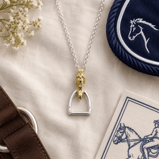 Silver and gold equestrian stirrup pendant necklace on a beige surface with floral and fabric accents.