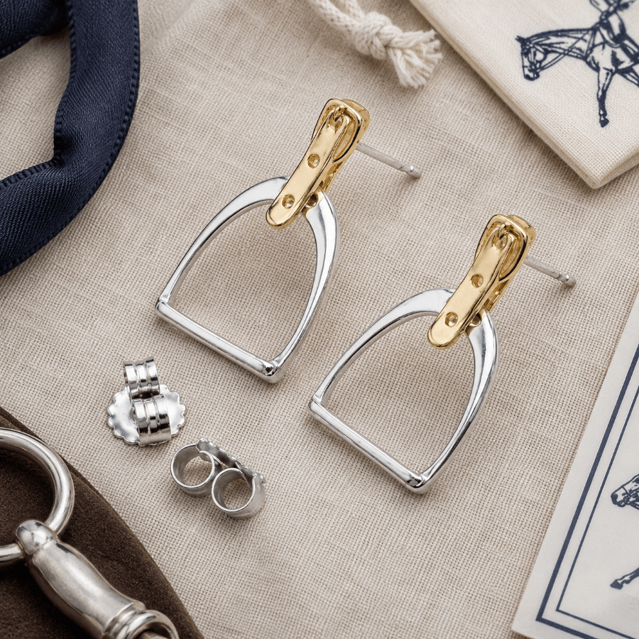 Pair of gold and silver stirrup earrings on a textured surface with other equestrian-themed items.