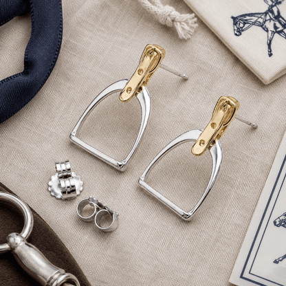 Pair of gold and silver stirrup earrings on a textured surface with other equestrian-themed items.