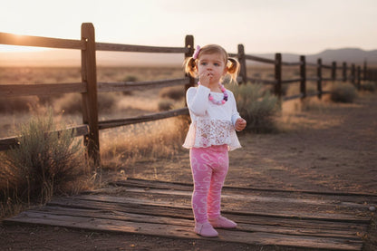 Young girl wearing a white top with lace detail and pink pants on a split rail fence background