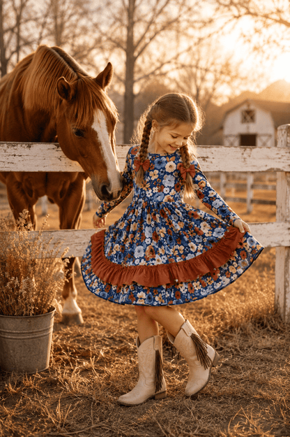 A young girl in a blue floral long-sleeve dress with a brown ruffle hem and beige cowboy boots stands beside a white wooden fence at a sunny farm, touching the head of a brown horse. A barn and dry grass are visible in the background.