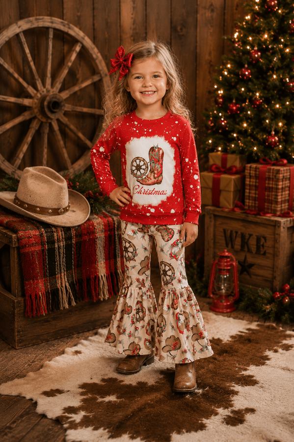 A young girl stands in a rustic Christmas-themed room wearing a red long-sleeve top with a cowboy boot graphic and white flared pants with a cowboy print. She has a red bow in her hair, and the background features a decorated Christmas tree, wooden wagon wheel, and wrapped gifts.