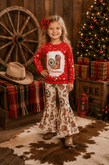 A young girl stands in a rustic Christmas-themed room wearing a red long-sleeve top with a cowboy boot graphic and white flared pants with a cowboy print. She has a red bow in her hair, and the background features a decorated Christmas tree, wooden wagon wheel, and wrapped gifts.
