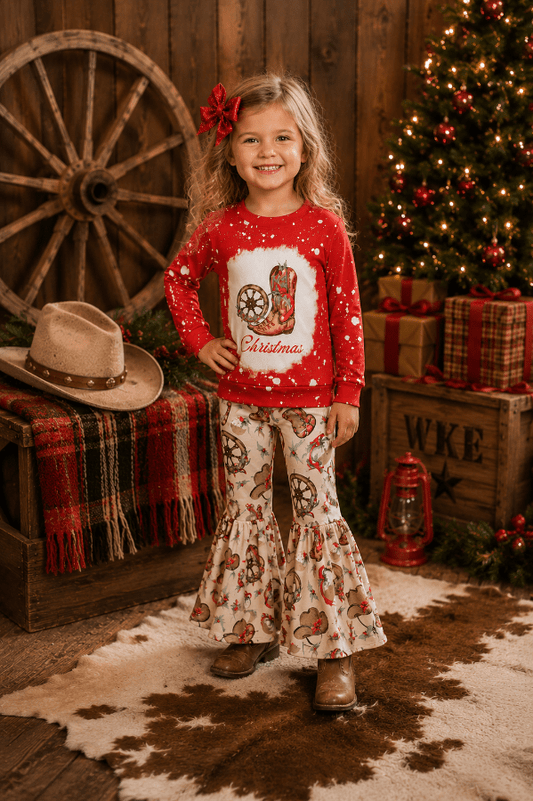 A young girl stands in a rustic Christmas-themed room wearing a red long-sleeve top with a cowboy boot graphic and white flared pants with a cowboy print. She has a red bow in her hair, and the background features a decorated Christmas tree, wooden wagon wheel, and wrapped gifts.