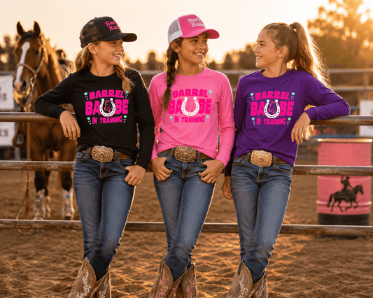 Three young women stand in a horse arena wearing matching long-sleeve graphic shirts with 'BARREL BASH' and a horseshoe design. The shirts are in black, pink, and purple, paired with jeans, western boots, and hats, with a horse and barrel visible in the background during golden hour.
