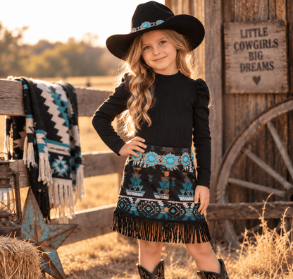 A young girl stands outdoors in a rustic barn setting wearing a black long-sleeve top, a black fringed skirt with turquoise and white geometric patterns, a black cowboy hat, and cowboy boots. A wooden fence, wagon wheel, and sign reading 'LITTLE COWGIRLS BIG DREAMS' are visible in the background.