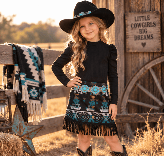 A young girl stands outdoors in a rustic barn setting wearing a black long-sleeve top, a black fringed skirt with turquoise and white geometric patterns, a black cowboy hat, and cowboy boots. A wooden fence, wagon wheel, and sign reading 'LITTLE COWGIRLS BIG DREAMS' are visible in the background.