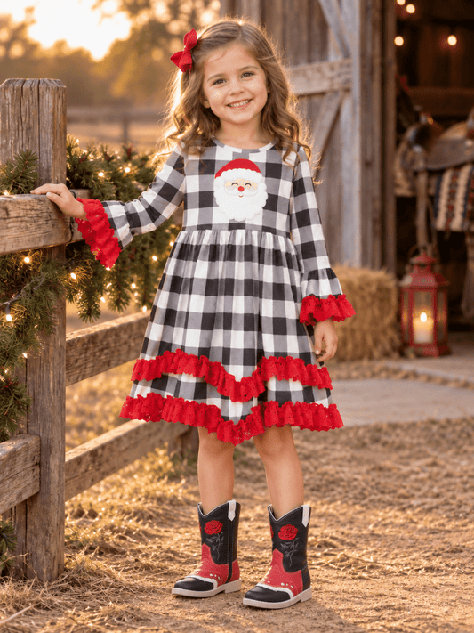 A young girl stands on a rustic barn porch wearing a black and white buffalo plaid dress with red ruffle trim and a Santa Claus face applique on the chest. She is wearing red and black cowboy boots and has a red bow in her hair.