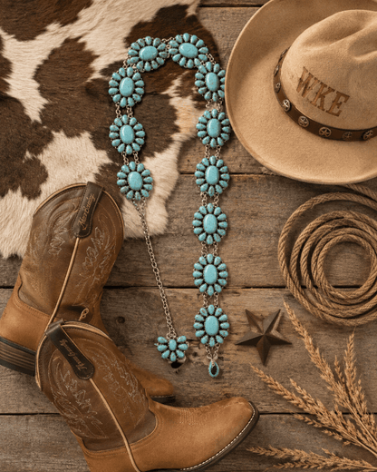 A turquoise and silver beaded necklace is arranged on a rustic wooden surface with cowboy boots, a tan hat, rope, star, and dried wheat. The necklace is the apparent product, shown in a styled flat-lay with western props.