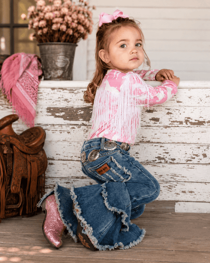 A young girl kneeling on a rustic wooden porch wearing a pink and white patterned long-sleeve fringed top, blue denim flare jeans with frayed hems, and pink glittery cowboy boots. She has a pink bow in her hair, with a wooden saddle and a bucket of pink flowers in the background.