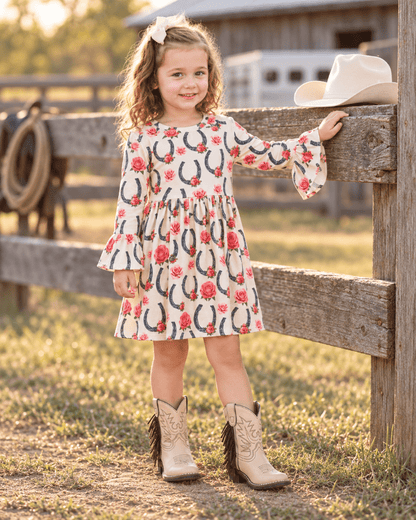 A young girl stands outdoors in front of a wooden fence wearing a cream long-sleeve dress with a red and black horseshoe and rose print, paired with tan western boots. A white cowboy hat rests on the fence beside her.