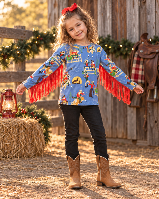 A young girl stands in a rustic barn setting wearing a blue long-sleeve shirt with cowboy-themed graphics and bright red fringe on the sleeves, paired with black pants and brown cowboy boots.