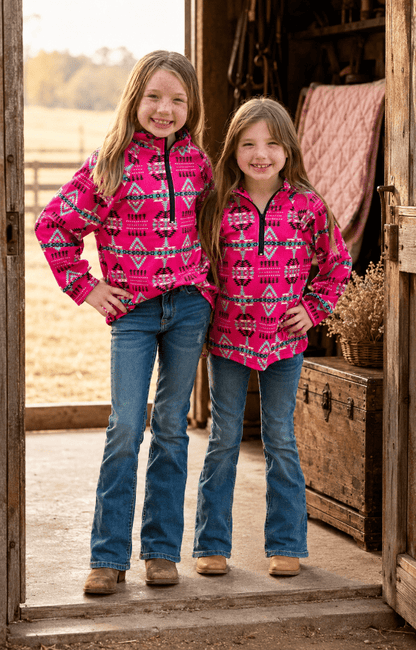 Two young girls standing in a rustic barn doorway, wearing matching bright pink quarter-zip fleece pullovers with a multicolor geometric pattern and blue jeans.