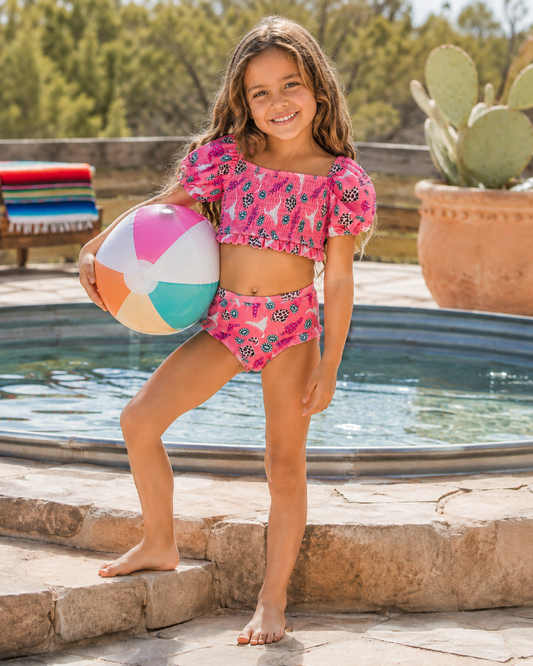 A young girl stands barefoot on stone steps beside a swimming pool, holding a colorful beach ball. She is wearing a bright pink two-piece swimsuit with a floral and butterfly print.