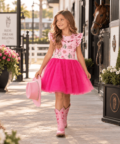 A young girl wearing a pink and white floral print top with a bright pink tulle skirt and pink cowboy boots walks down a sunny outdoor stable walkway, holding a pink hat.