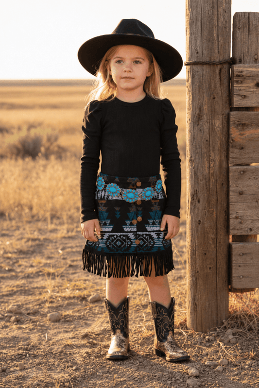 A young girl stands outdoors in a rural setting next to a wooden post, wearing a black long-sleeve top, a black fringed skirt with a colorful turquoise and black patterned waistband, and black cowboy boots.