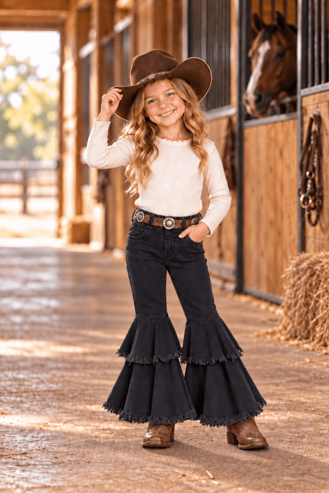 A young girl stands in a horse stable wearing a brown cowboy hat, white long-sleeve top, dark flared jeans with ruffled hems, a western belt, and brown boots. A horse is visible in the background.