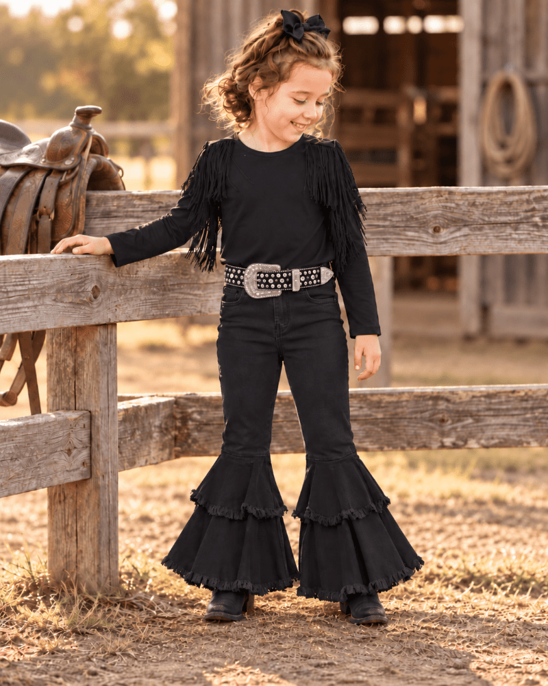 A young girl stands outdoors in front of a rustic wooden fence and barn, wearing a black long-sleeve top with fringe, black flared pants with a tiered ruffle hem, a silver studded belt, and black boots.