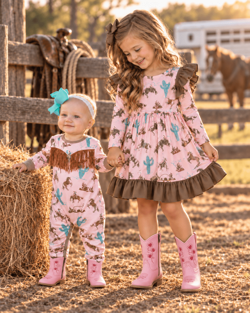 Two young girls in matching pink western-themed outfits standing on a sunny ranch. The older girl wears a long-sleeve pink dress with a brown ruffled hem and pink cowboy boots, while the younger girl wears a matching pink one-piece jumpsuit with brown fringe and pink boots.