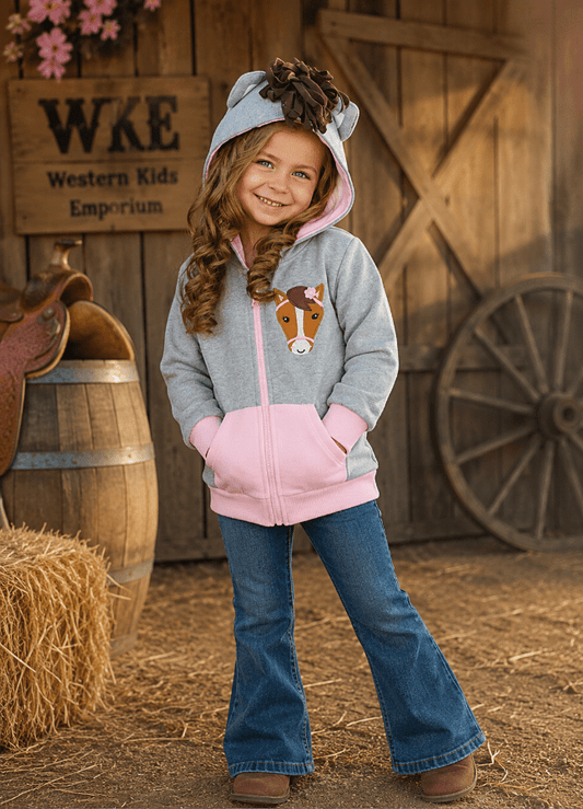 A young girl stands in a rustic barn setting wearing a light gray and pink zip-up hoodie with a horse graphic on the front, blue jeans, and brown boots. The background features a wooden barrel, hay bale, wagon wheel, and a wooden sign that reads 'WKE Western Kids Emporium'.