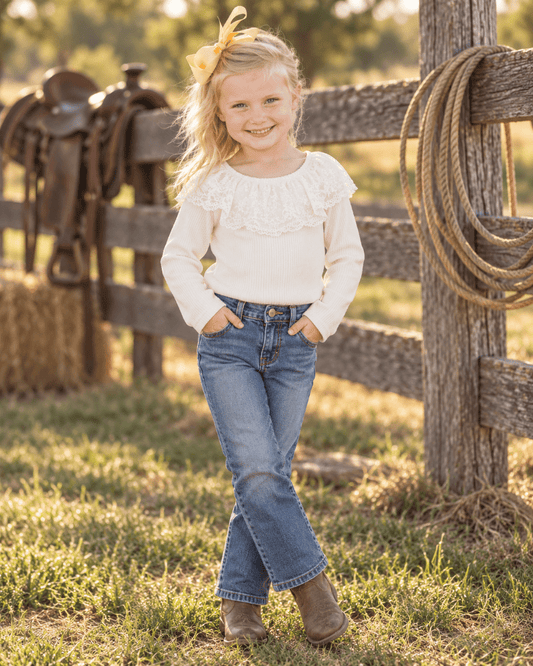 A young girl stands outdoors in a sunny ranch setting, wearing a cream long-sleeve ruffle-trim top, blue jeans, and brown boots. A wooden fence, rope, and saddles are visible in the background.