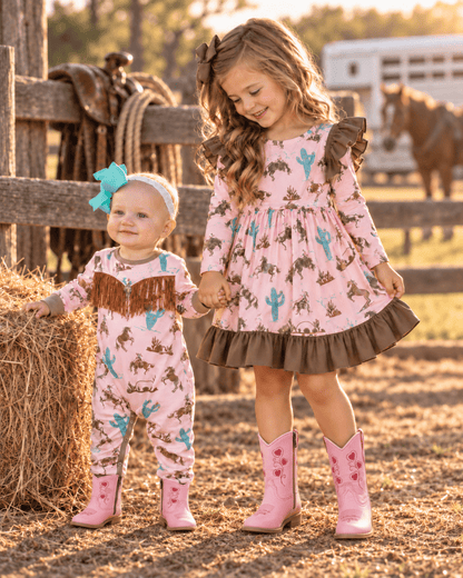 Two young girls in matching pink western-themed outfits standing on a sunny ranch. The older girl wears a long-sleeve pink dress with a brown ruffled hem and pink cowboy boots, while the younger girl wears a matching pink one-piece jumpsuit with brown fringe and pink boots.