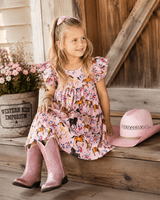 A young girl sits on rustic wooden porch steps wearing a pink flutter-sleeve dress with a multicolor horse print, pink glitter cowboy boots, and a pink hat rests beside her. A potted flower arrangement and a wooden barn wall are visible in the background.