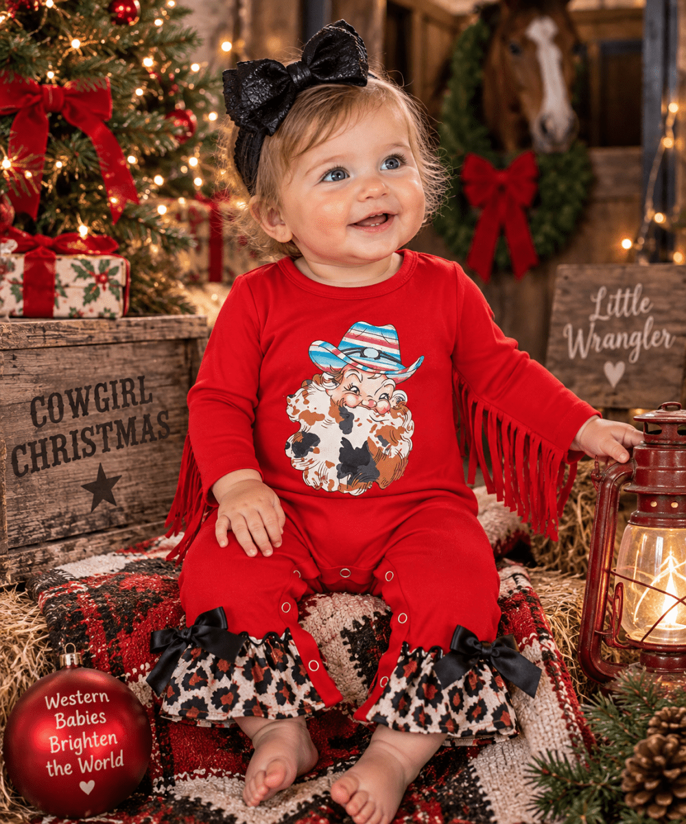 A toddler girl wearing a red long-sleeve romper with fringe, leopard-print ruffle hems, and a Santa graphic on the front, sitting in a rustic Christmas-themed setting with a tree, lantern, and wooden crates.